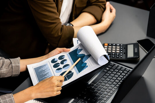 Cropped working asian smart manager with coworker discussing paperwork comparing business data report on laptop at modern home office, taking note with customer segment to marketing plan. Infobahn.