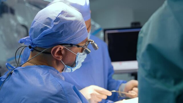 Male Caucasian doctor in cap, mask and surgical loupes working at surgery. Side view portrait of a professional neurosurgeon working in a team.