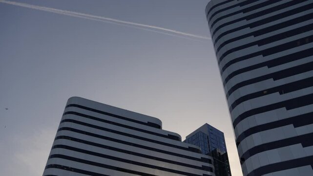 Low angle view of modern high-rise buildings with smooth curved lines. Contrail from airplane crosses the sky at dusk. Driving under the bridge. Stylish cityscape. 