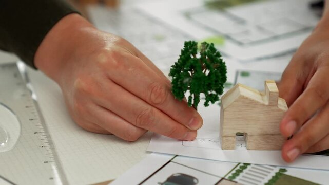 A video of architect using tree and home model to measure blueprint. Architect designing house construction on a table at studio, with architectural plan scattered around. Focus on hand. Delineation.