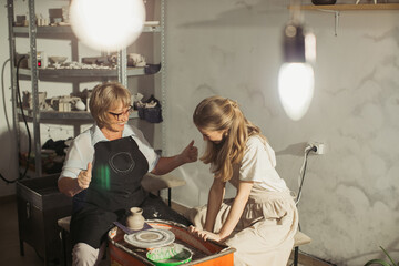 Senior potter showing thumbs up to young apprentice working on pottery wheel
