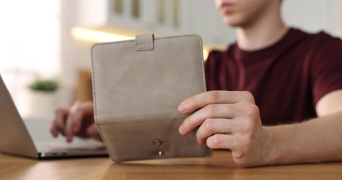 Man with passport using laptop at wooden table indoors, closeup