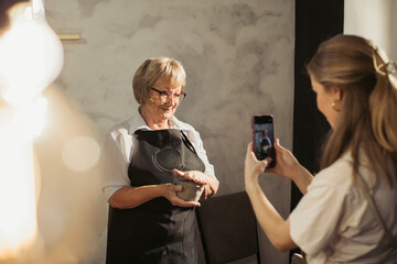 Senior woman showing pottery while student is taking pictures with smartphone