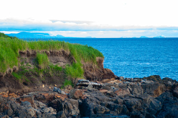A rugged coastal cliff covered in green grass overlooks a rocky shoreline and a calm blue sea, with...