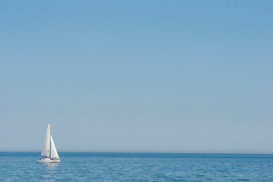 A single white sailboat gracefully glides across the calm, blue expanse of the sea under a clear, vast sky. Perfect for themes of freedom, tranquility, travel, and adventure on water