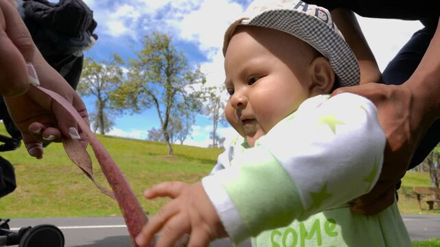Adorable baby wearing a hat and romper learns to walk in a park, reaching for a leaf held by their parent