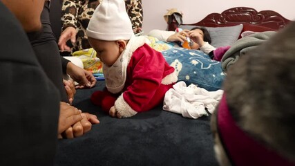 Adorable baby boy wearing a Santa Claus costume, playing on a bed surrounded by family and a Chihuahua