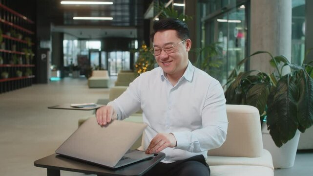 Chinese young businessman closing laptop computer after finishing work at hotel lobby. Smiling manager busy working online, remote job. E-learning, browsing internet on netbook looking at camera.