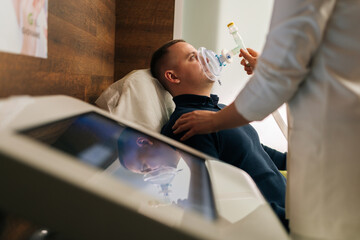 Side view of unrecognizable female doctor assisting to male patient undergoing respiratory training with oxygen mask, providing essential care and monitoring progress in modern medical clinic.