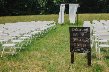 Rustic outdoor wedding ceremony seating with white folding chairs, wooden “pick a seat” sign...