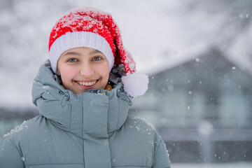 Young caucasian girl model with happy smile wearing a red santa hat during a winter snowfall holiday outdoors with copy space.