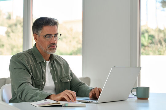 Busy serious older mature middle aged man wearing glasses looking at computer tech sitting at home table, using laptop hybrid working online, elearning, browsing web, searching online in living room.