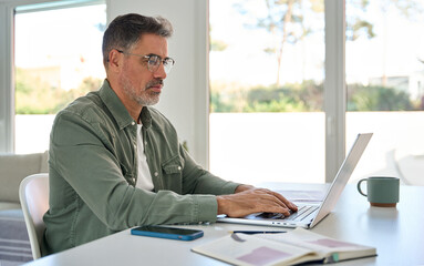 Busy older mature middle aged man wearing eyeglasses looking at computer technology sitting at...