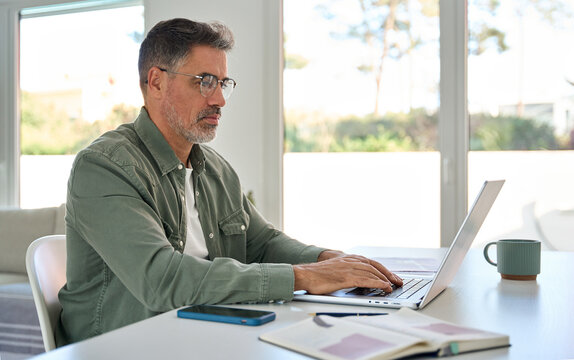 Busy older mature middle aged man wearing eyeglasses looking at computer technology sitting at table, using laptop hybrid working online, elearning, browsing web, searching online at home.