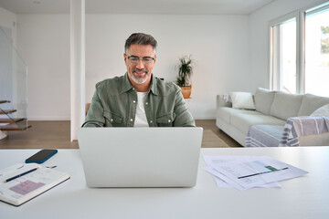 Obraz na płótnie Canvas Smiling happy older mature middle aged professional man wearing eyeglasses looking at computer sitting at living room table, using laptop hybrid working, paying bills, browsing online at casual home.
