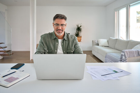 Smiling happy older mature middle aged professional man wearing eyeglasses looking at computer sitting at living room table, using laptop hybrid working, paying bills, browsing online at casual home. - Powered by Adobe