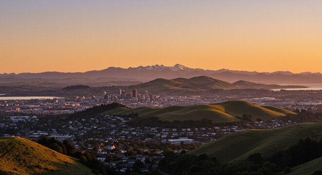 Cityscape view from rolling hills at sunset with distant mountains and orange sky above the horizon - Powered by Adobe