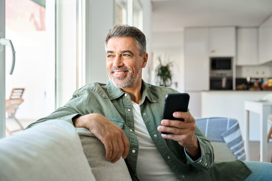 Relaxed happy mature middle aged man holding cell mobile phone using smartphone sitting at home on sofa, relaxing scrolling social media, checking financial app, buying online, looking away at window.
