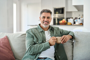 Portrait of happy middle aged man holding coffee cup relaxing on couch at home. Smiling mature older man drinking tea looking at camera sitting on cozy sofa chilling in modern kitchen living room.