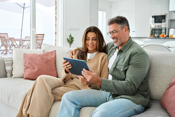 Middle aged man and woman hugging sitting on sofa at home together relaxing on couch using digital tablet computer. Happy mature old couple buying online browsing internet in modern house living room.