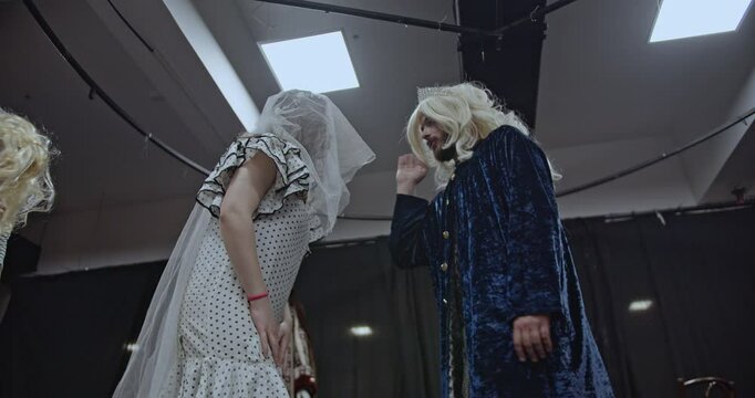 Actors rehearse a scene on a theater stage. A veiled woman hands a phone to a person with a cane while a king looks on.