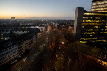 A view of the Cologne city skyline at dawn, featuring a tall office building with glowing windows, a city street, and white and red traffic lights.