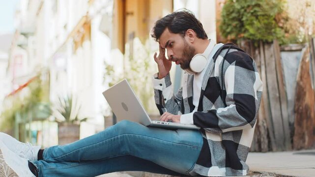 Displeased Indian man using laptop typing browsing working, loses becoming surprised sudden lottery results bad news fortune loss game fail computer virus. Arabian guy sitting on urban city street.
