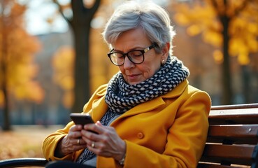 Elderly woman in autumn park sits on bench uses smartphone. Senior female in eyeglasses, yellow coat, checkered scarf, reads news or texts. Mature lady uses mobile device outdoors.