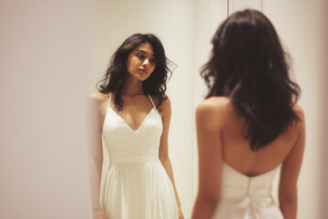 Fototapeta premium A young Asian woman with long black hair stands in front of a mirror, wearing a white dress. She gazes thoughtfully at her reflection in a softly lit room.