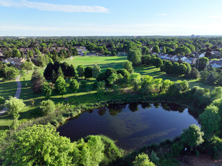 A serene pond reflects the surrounding lush greenery of a park and residential neighborhood under a...
