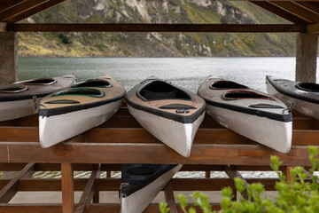 Beneath a wooden shelter by the lake, colorful kayaks lie quietly, waiting. The stillness suggests...