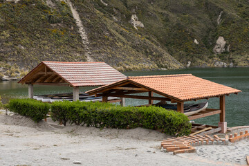 Two wooden shelters with red tiled roofs stand by a serene lake, nestled among green mountains. A peaceful spot for resting, storing boats, and enjoying the natural scenery.

