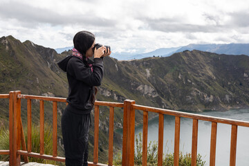 A woman stands with her camera on a wooden lookout, focused on photographing the breathtaking Quilotoa Lake below. The scene evokes creativity, tranquility, and admiration for nature.


