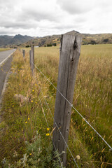 From a moving car, a wooden fence post with barbed wire marks the boundary of a rural field. The...