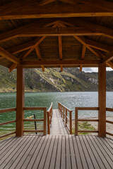 A captivating view from inside a wooden pier reveals intricate timber construction alongside a breathtaking panorama of the calm lake and lush mountains beyond.

