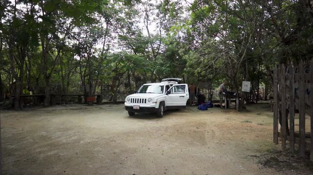 A white car arrives at a wooded parking area near a Cenote in Yucatan, Mexico. People gather their gear, ready to explore the underground caves and crystal-clear waters.