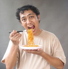 Portrait of handsome asian man eat instant noodle on grey background. He's feel surprise and...