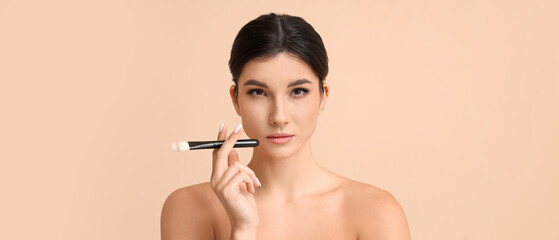 Beautiful young woman with makeup brush on beige background