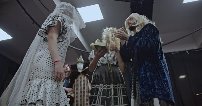 Actors rehearse a scene on a theater stage. They wear costumes and wigs and interact with props.