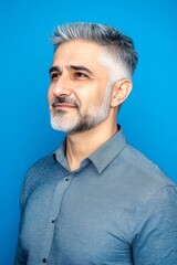Fototapeta premium Blue solid background. A graceful mature man with a modern hairstyle. Trendy men's curtain bangs haircut - midlife middle eastern man with short hair. Creative of style for aging men.
