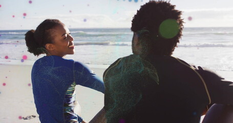 Sitting wetsuit-clad adult couple watching gentle ocean waves lapping shore at beach, sunny sky