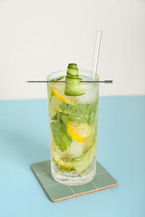 Glass of mojito with lemon, cucumber and ice cubes on blue table near white wall
