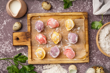 Wooden board with colorful raw dumplings and flour on dark background