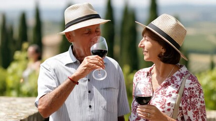 Elderly couple enjoying red wine outdoors in vineyard, wearing summer hats and casual clothing, surrounded by green landscape and cypress trees, smiling and relaxed in warm sunlight, joyful atmosphere - Powered by Adobe