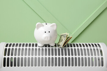 Piggy bank with money on electric convector heater near green wall at home. Heating saving concept