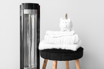 Electric heater and piggy bank with clothes on stool against white background. Heating saving...