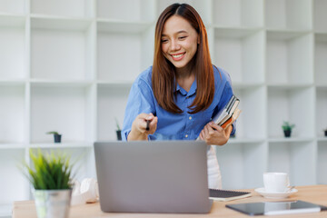 Smiling young asian woman sitting at desk working on laptop taking notes in notebook, happy...