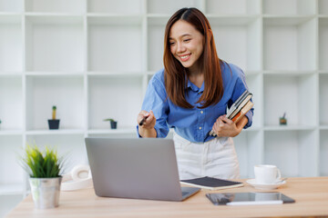 Smiling young asian woman sitting at desk working on laptop taking notes in notebook, happy...
