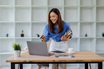 Smiling young asian woman sitting at desk working on laptop taking notes in notebook, happy...