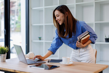 Smiling young asian woman sitting at desk working on laptop taking notes in notebook, happy...